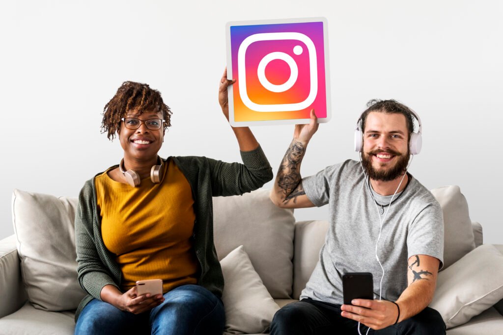 Duas pessoas sorrindo sentadas em um sofá com fones de ouvido e smartphones, segurando um ícone do Instagram.